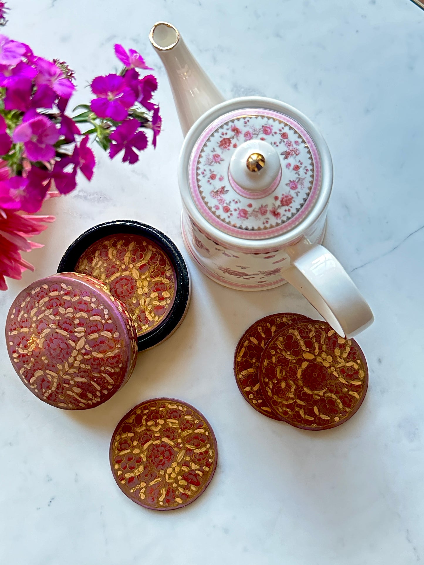 a set of beautiful artisanal hand-painted pink and gold coasters kept on a table with some pink flowers and a kettle