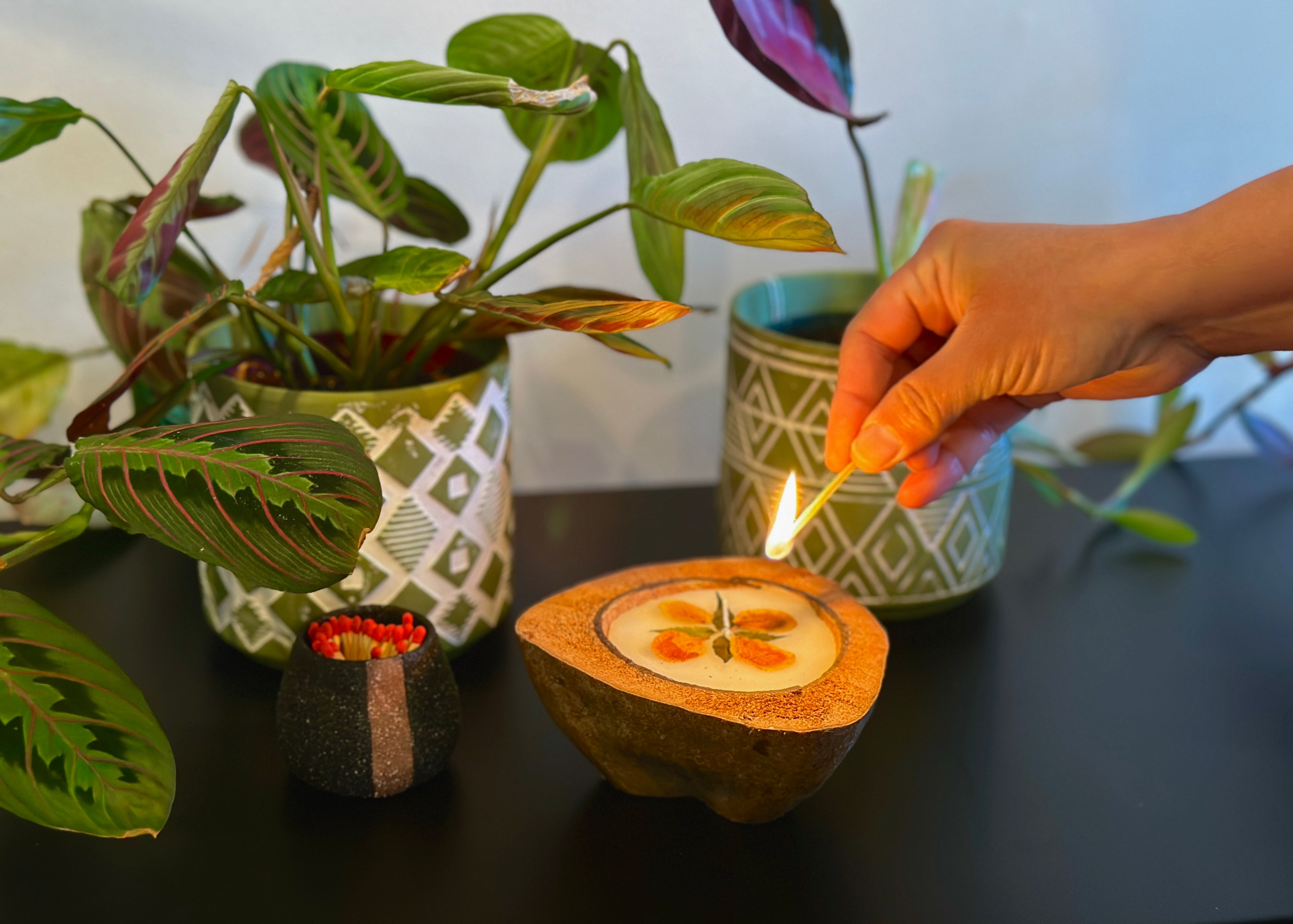 a hand using a matchstick to light a handmade coconut candle kept in a beautiful table setting with plants