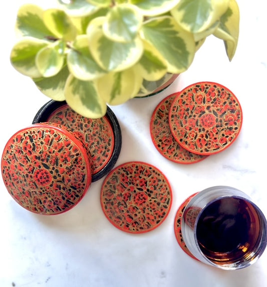 a set of beautiful artisanal hand-painted red and gold coasters kept on a table with a plant and a glass of wine